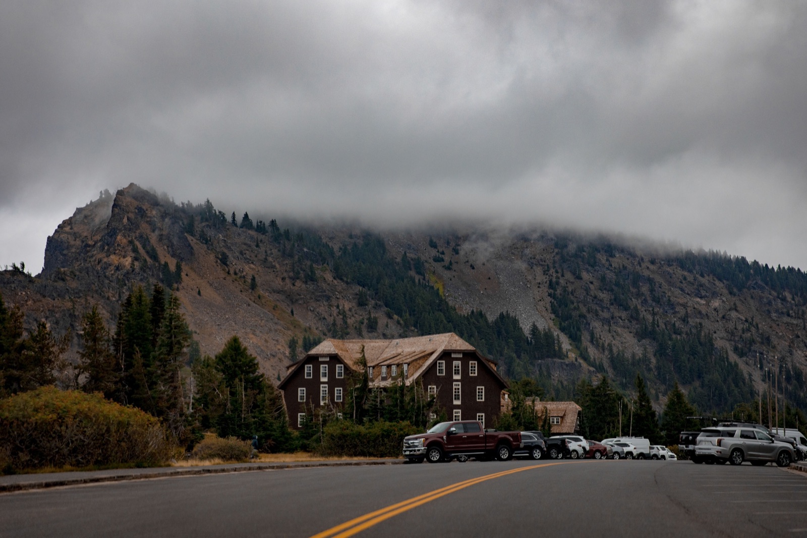 Crater Lake Lodge under low cloud, with a forested ridge behind and cars parked along the road