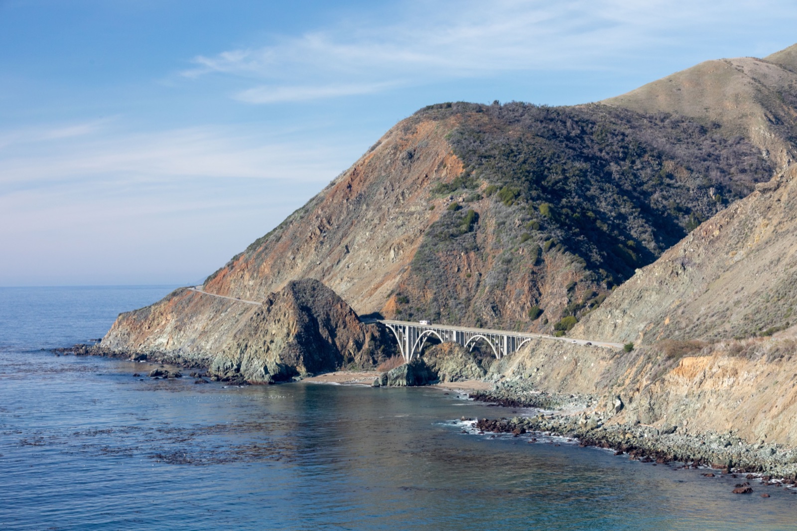 Highway 1 crossing the Big Creek Bridge below steep brown hills on the Big Sur coast, with calm blue ocean in the foreground