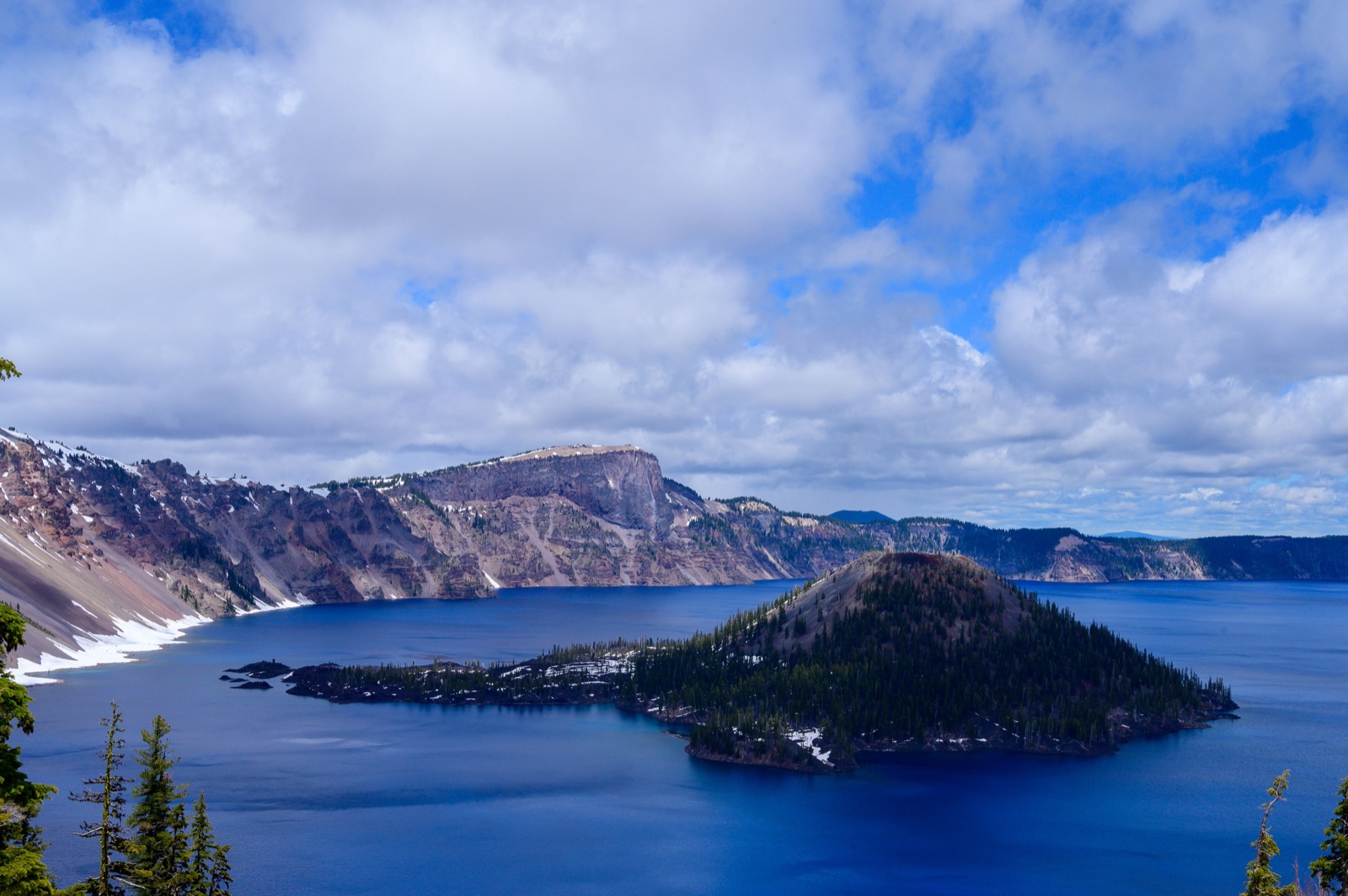 Wizard Island rising from the deep blue water of Crater Lake, with snow-streaked caldera walls and clouds behind