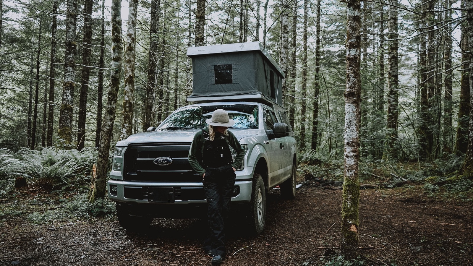 A person in a wide-brimmed hat leaning on a pickup truck with a pop-up rooftop camper, parked among mossy conifers and ferns
