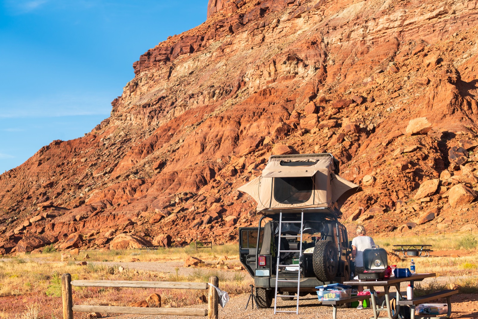A Jeep with a rooftop tent parked at a campsite below red sandstone cliffs in Utah