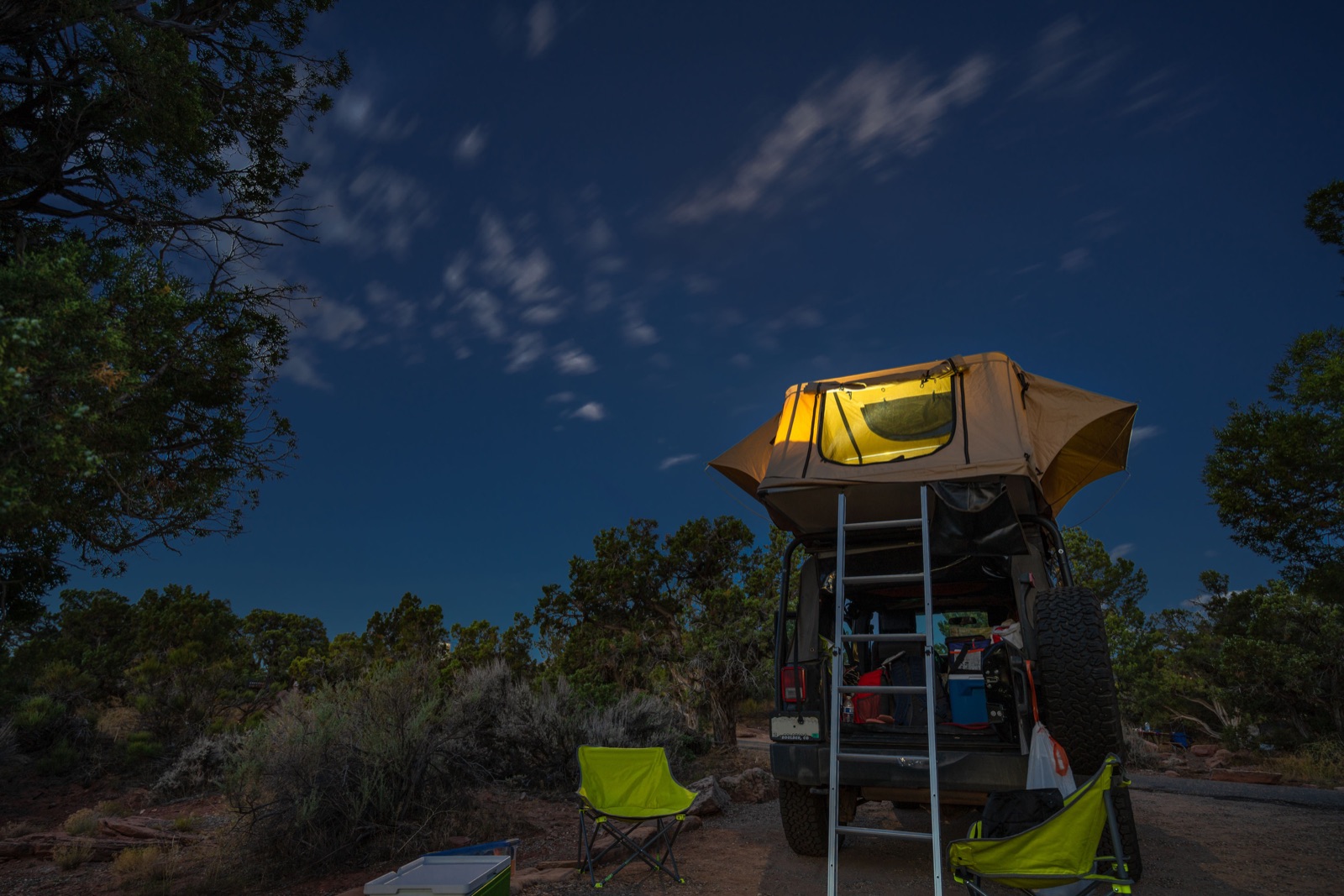 A rooftop tent lit up at night on a vehicle parked among junipers near Moab