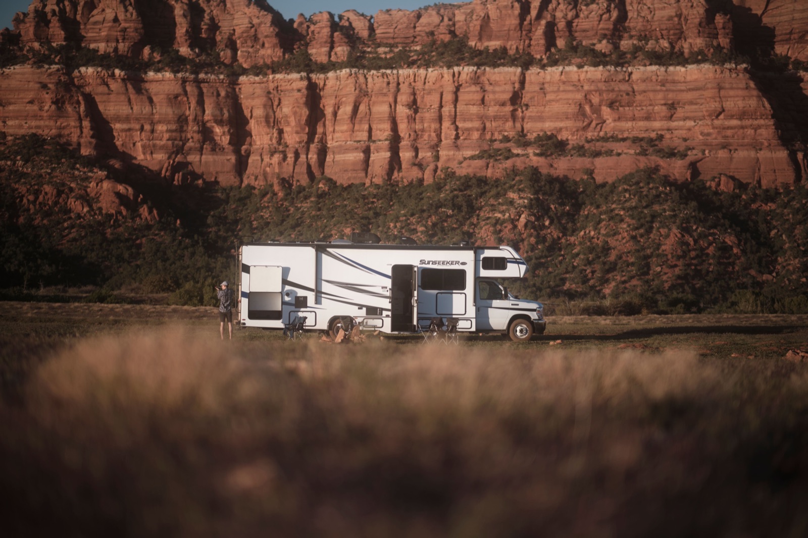A Class C motorhome parked on open ground below red sandstone cliffs