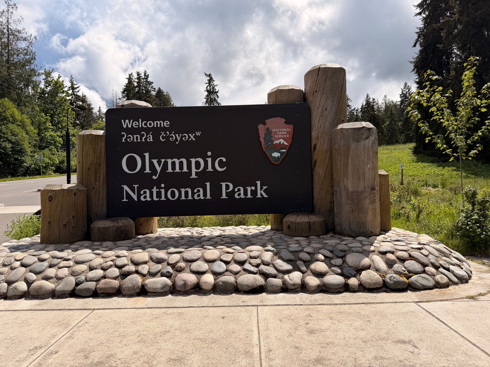 The Olympic National Park welcome sign beside a path and a stone border on a sunny day