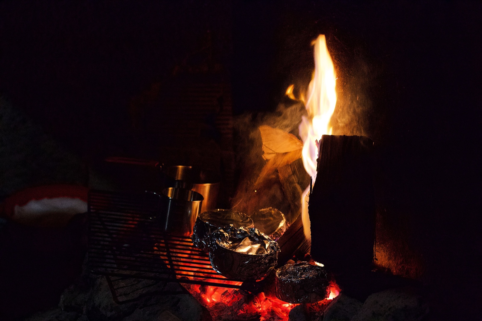 Foil-wrapped food on a grill grate next to a burning campfire at night