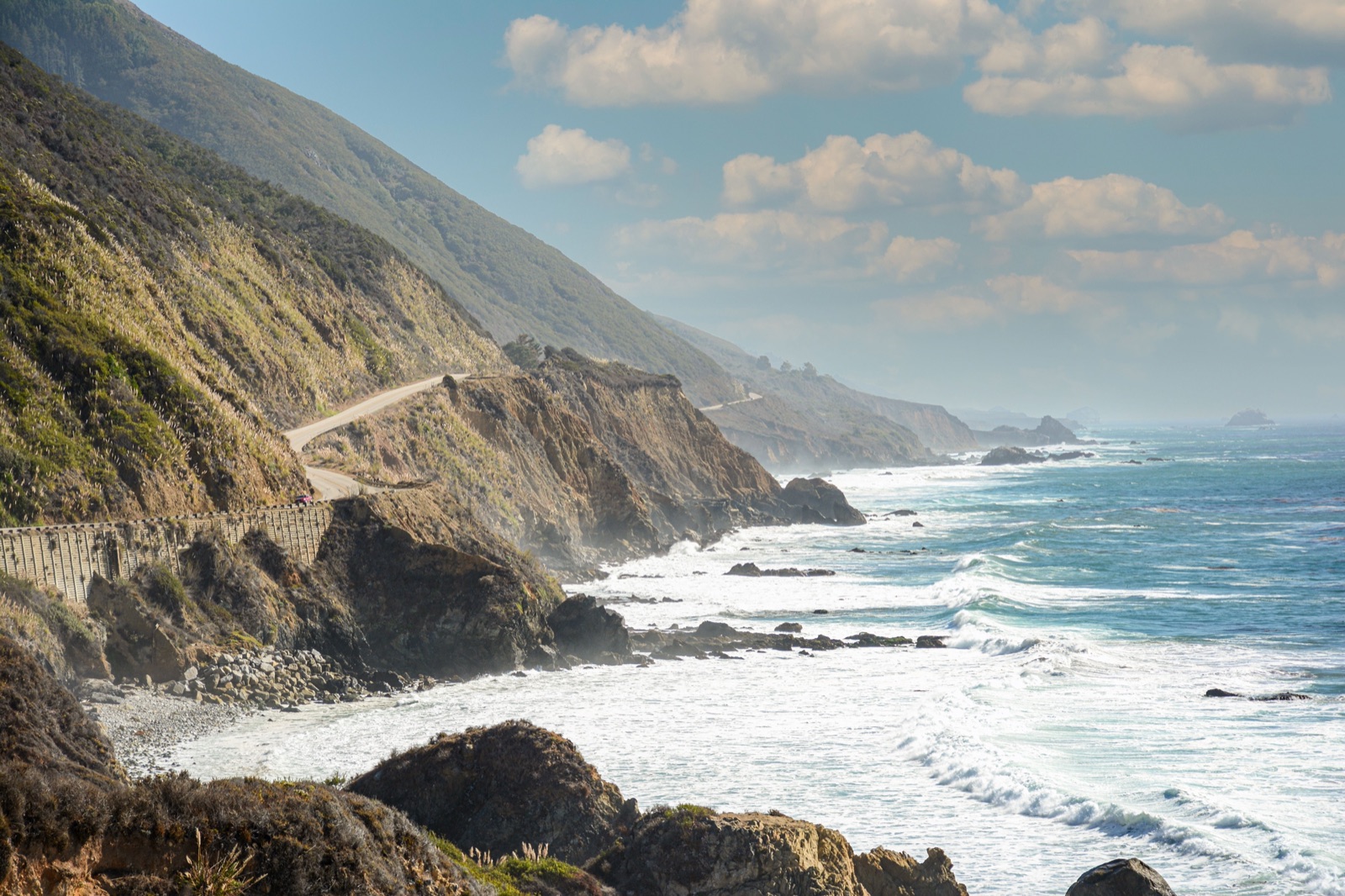 A coastal highway winding along steep green cliffs above white surf on the California coast