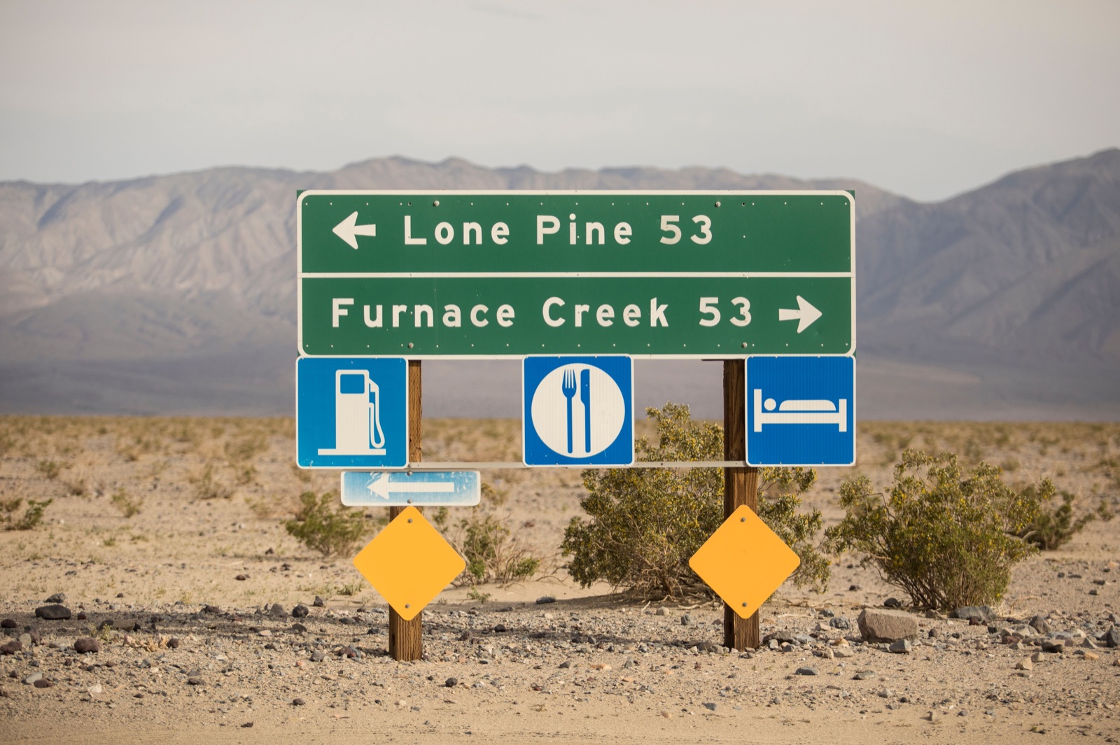 Green road sign in the Death Valley desert pointing to Lone Pine and Furnace Creek, 53 miles each way, with gas, food and lodging symbols