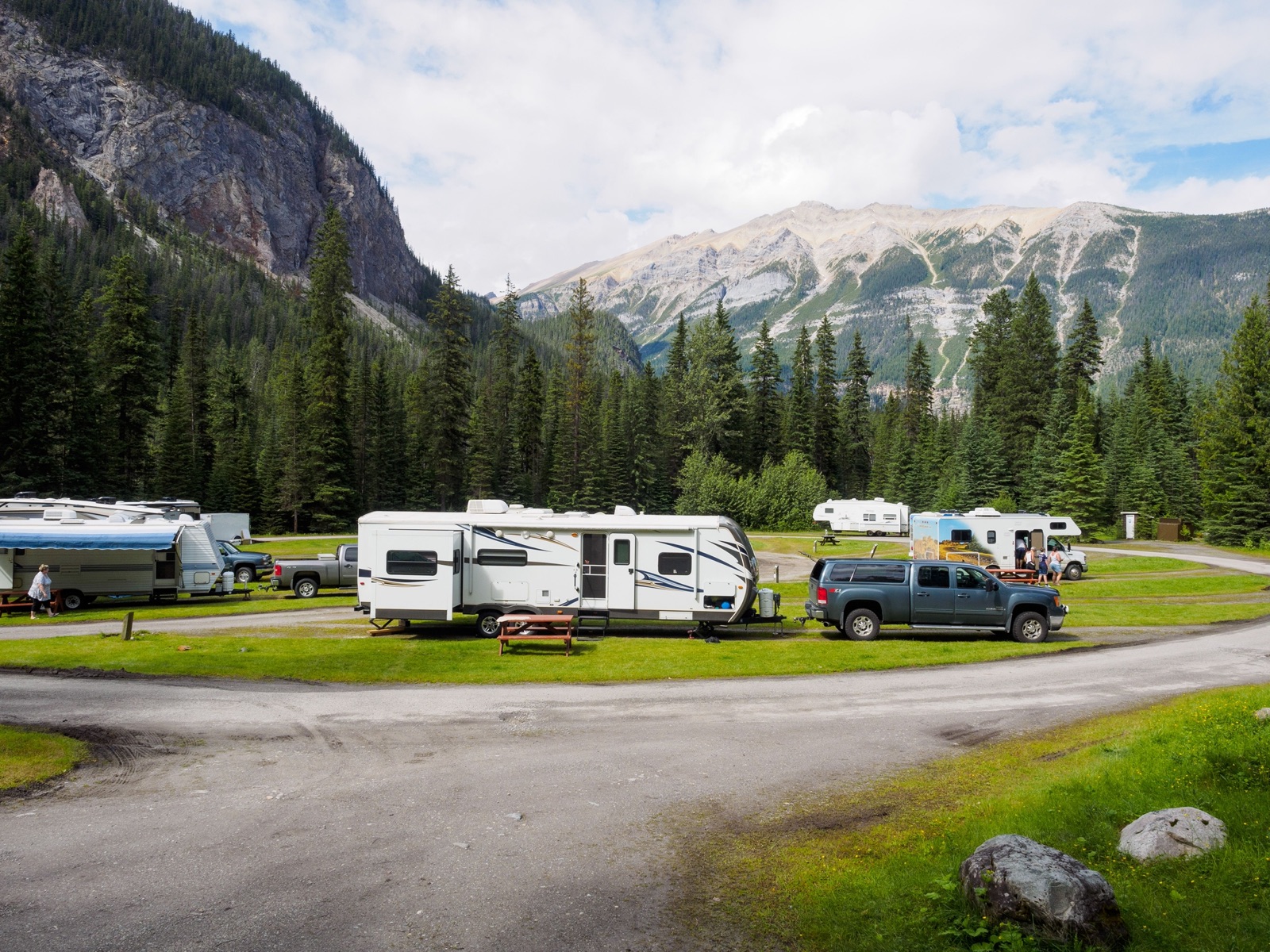 Travel trailers and pickups parked at a grassy campground below forested mountains